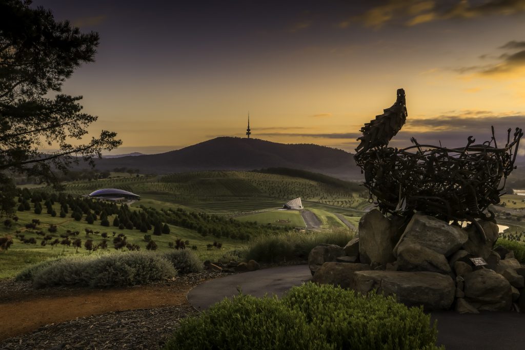A large metal sculpture of a bird in a nest overlooks rolling green hills at sunset. In the distance, a mountain with a communications tower rises beneath a glowing orange and purple sky.
