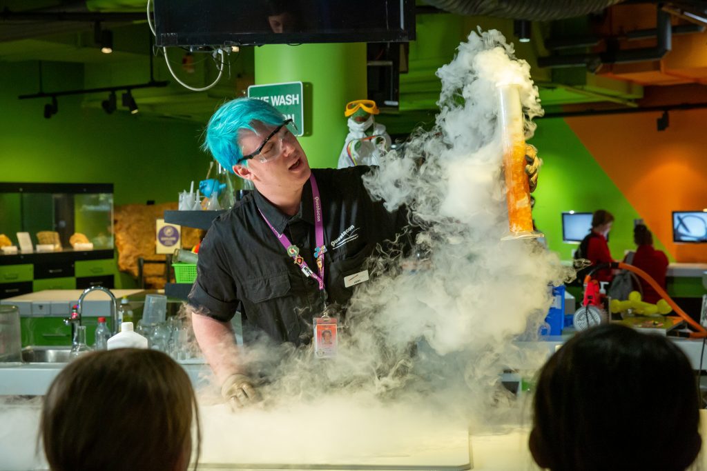 A science educator wearing safety glasses demonstrates a chemistry experiment in a laboratory setting, holding a tall container filled with orange material while thick white vapour rises around the bench. 

