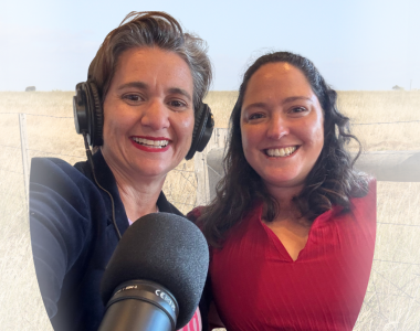 Portrait-oriented image of two people seated side by side at a table with a professional podcast microphone in the foreground. One person is wearing studio headphones, and both are positioned in front of a plain wall with exposed brick visible to one side.