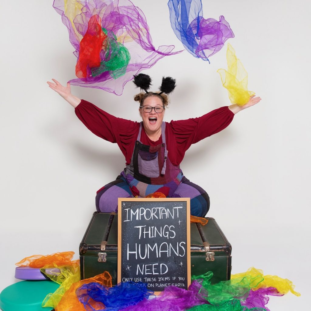 A person wearing a red top, purple patchwork overalls, glasses, and a headband with black pom-pom antennae sits on a large trunk in a studio setting. Colourful translucent scarves are suspended and scattered around the scene. A chalkboard sign on the trunk reads “Important Things Humans Need,” with smaller text underneath. The background is plain light grey.
