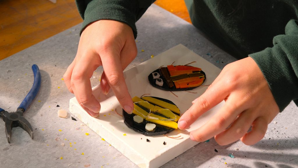 Hands carefully arrange colourful pieces of glass into a bee-shaped mosaic on a work surface. A pair of pliers and another completed bee design sit nearby.
