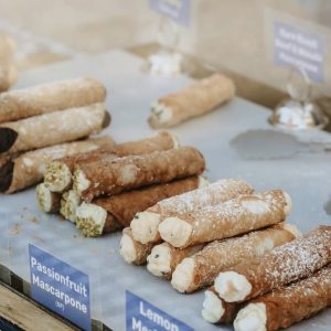 Assorted cannoli filled with cream and topped with powdered sugar, displayed in a food stall case with flavor labels including roasted pistachio ricotta and passionfruit mascarpone.