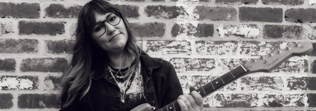 Performer holding an electric guitar in front of a brick wall in a black-and-white promotional portrait.