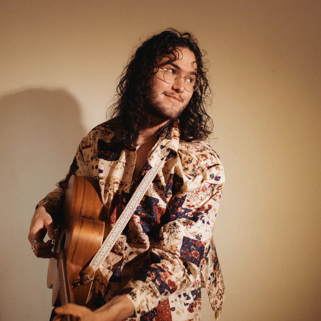 Promotional portrait of a musician holding an acoustic guitar and wearing a patterned shirt. Warm lighting casts a soft shadow on the neutral background.