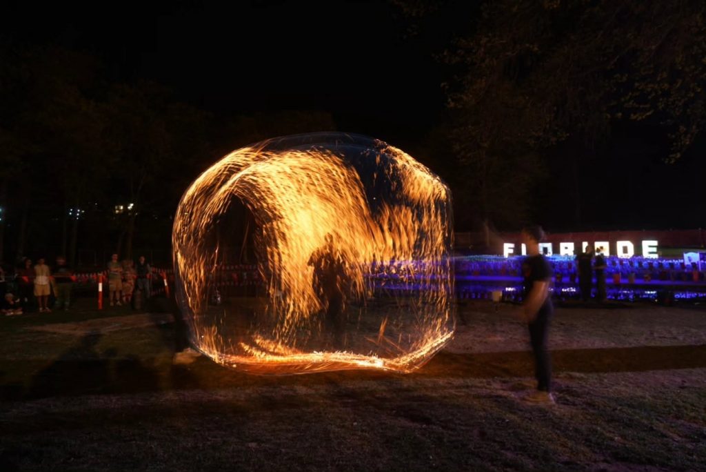Fire performer creating a circular trail of sparks at night, with illuminated Floriade signage in the background.