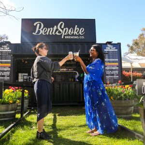 Two visitors raise drinks in a toast outside the BentSpoke Brewing Co. container bar, surrounded by colourful tulip displays at an outdoor festival.