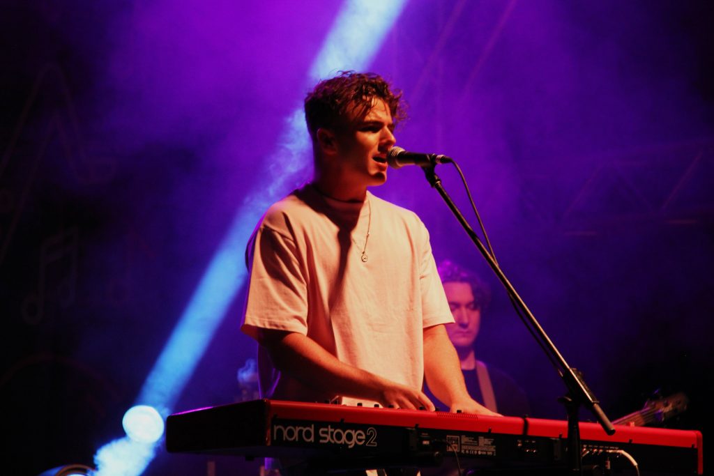 Performer singing into a microphone while playing a keyboard under purple stage lighting.