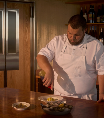 Head Chef Will Moyle, dressed in a white chef's jacket and apron, plates a dish at a wooden counter. Oysters, a lemon wedge and small dishes are arranged in the foreground, while wine bottles line shelves in the background.