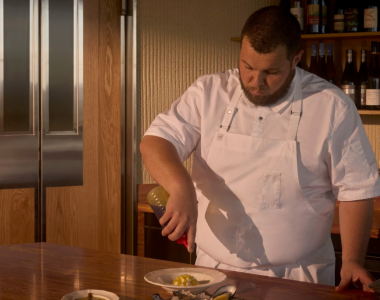 Head Chef Will Moyle, dressed in a white chef's jacket and apron, plates a dish at a wooden counter. Oysters, a lemon wedge and small dishes are arranged in the foreground, while wine bottles line shelves in the background.