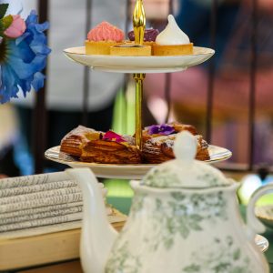 An elegant two-tiered stand displays assorted miniature pastries and sweets beside a floral teapot, creating a garden-inspired high tea setting.