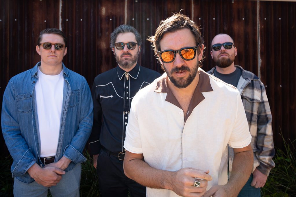 Four band members wearing sunglasses posing in front of a corrugated metal wall.