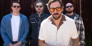 Four band members wearing sunglasses posing in front of a corrugated metal wall.