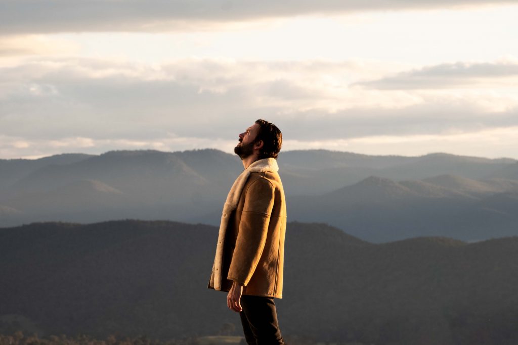 Performer standing on a mountain lookout with layered ranges and clouds in the background.