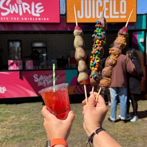 Hands holding three chocolate-coated fruit skewers and a red drink in front of a colorful Swirle and Juicelo kiosk on a sunny day.