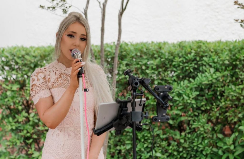 Performer singing into a microphone beside a tablet stand in an outdoor garden setting.