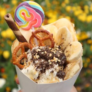 KusuKusu Dessert crepe filled with ice cream and banana slices, topped with chocolate cookie crumbs, chopped nuts, pretzels, a wafer stick, and a colorful lollipop.
