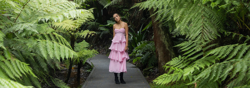 Performer standing on a boardwalk surrounded by large tree ferns and dense rainforest vegetation.