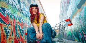 Promotional image of a person seated in a shopping trolley between two walls covered in colourful street art. The person wears a black helmet, a yellow shirt, and wide-leg blue jeans.