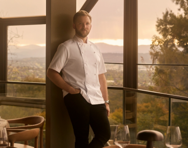 Lunetta Executive Chef Tristan Rebbettes stands beside floor-to-ceiling windows in a restaurant dining room, with set tables in the foreground and scenic views of trees and distant hills beyond.