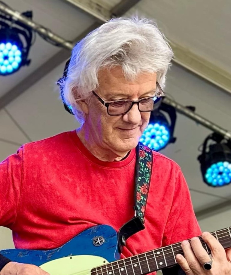 Performer playing an electric guitar on stage beneath blue concert lighting.