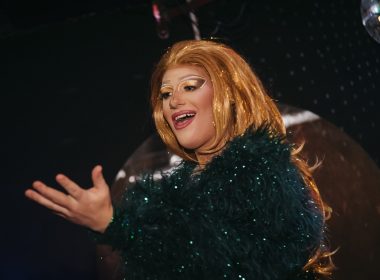 Promotional image of a stage performer wearing a glittering dark green feathered costume and a long blonde wig, illuminated by warm stage lighting against a dark backdrop.