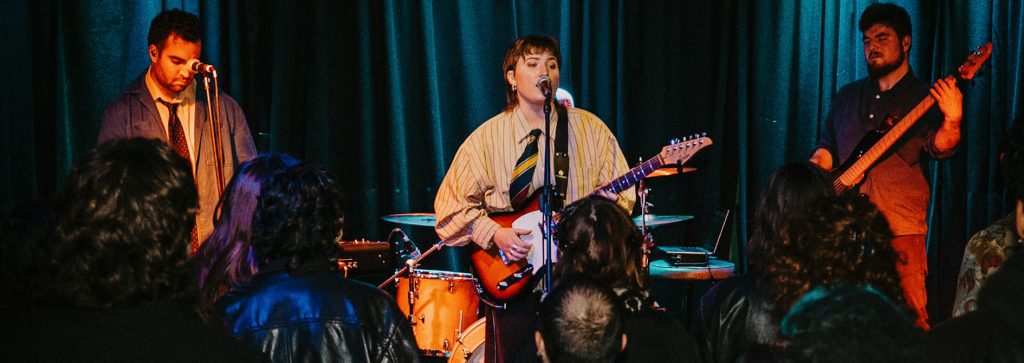 Band performing on stage with electric guitar, bass guitar and drums in front of an audience.