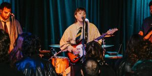 Band performing on stage with electric guitar, bass guitar and drums in front of an audience.
