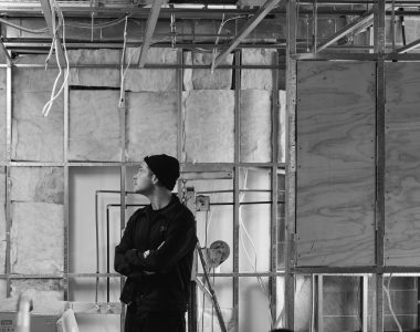 Black-and-white photograph of Head Chef Malcolm Hanslow standing inside a building under construction, looking upward at exposed wall framing, insulation, wiring and ceiling supports.