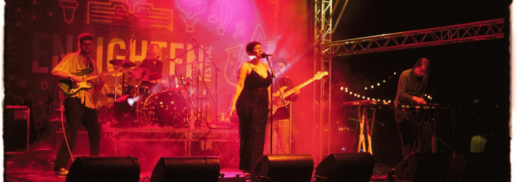 Band performing on an outdoor festival stage with vocals, guitars, keyboards and drums under coloured lighting.