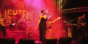 Band performing on an outdoor festival stage with vocals, guitars, keyboards and drums under coloured lighting.