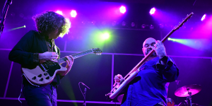Two guitarists performing on stage under purple and green concert lighting.