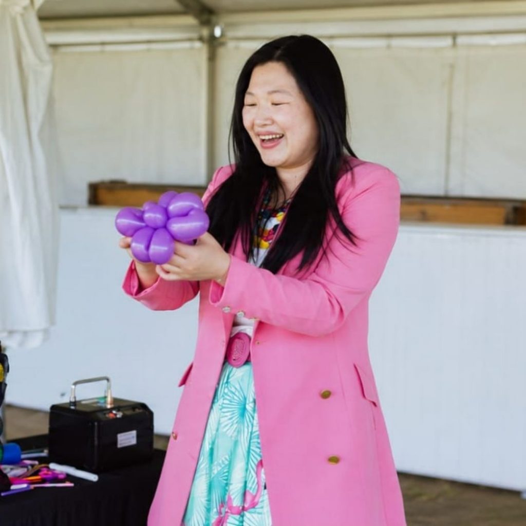 A performer stands inside a large event tent demonstrating balloon twisting, holding a purple balloon sculpture beside a table filled with colourful balloons and equipment.