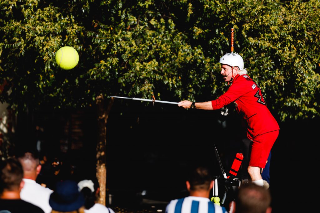 A street performer in a bright red costume and white helmet balances on a platform while extending a long rod toward a large tennis ball suspended in the air. Audience members watch from the foreground, with leafy trees forming the background.