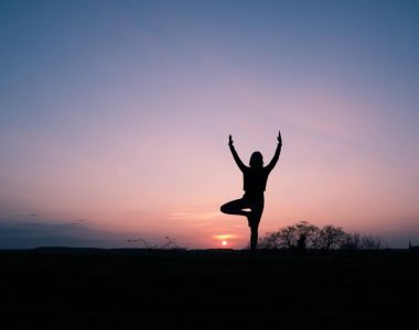 Silhouette of a person balancing in a yoga tree pose at sunset, standing against a pink and blue twilight sky with the sun low on the horizon.