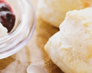 Close-up of two golden scones on a glass plate with a small glass bowl containing cream and dark berry jam. The image is softly lit with a warm, inviting appearance.