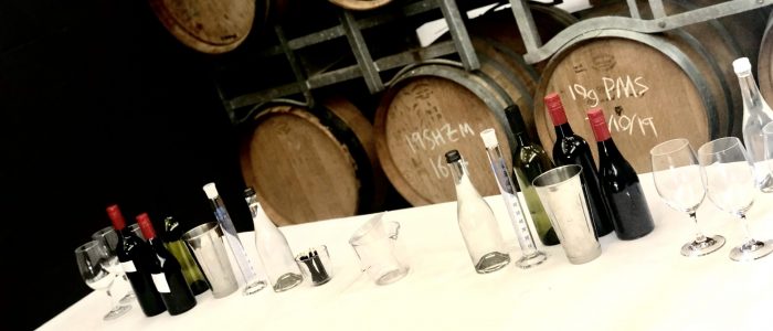 Wine tasting and measuring equipment arranged on a table in front of stacked wooden barrels in a winery cellar.