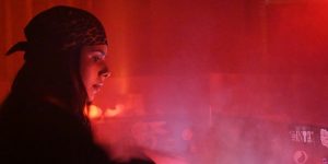 Woman in a bandana playing a dj set to a crowd. Thick red smoke is over the dance floor.